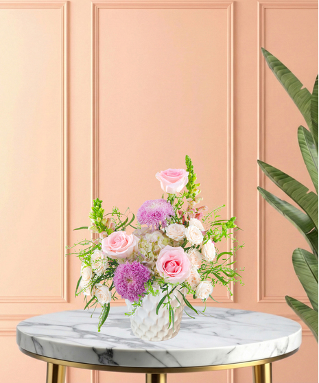Lavender Sunrise floral arrangement with peach and blush roses, lavender pom blooms, snapdragons, and hydrangeas in a white ceramic vase.