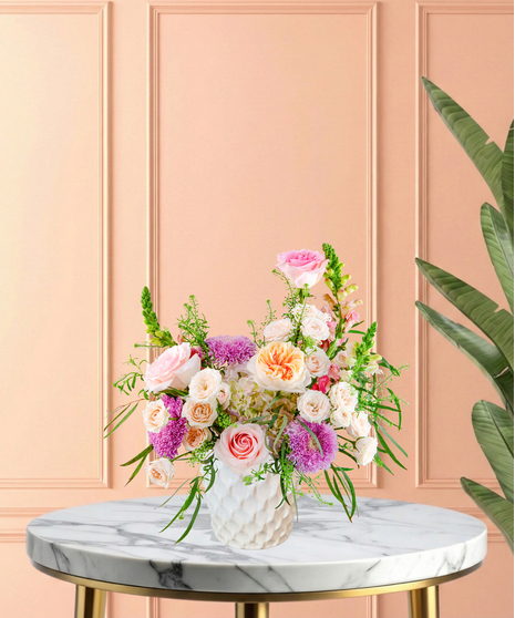 Lavender Sunrise floral arrangement with peach and blush roses, lavender pom blooms, snapdragons, and hydrangeas in a white ceramic vase.