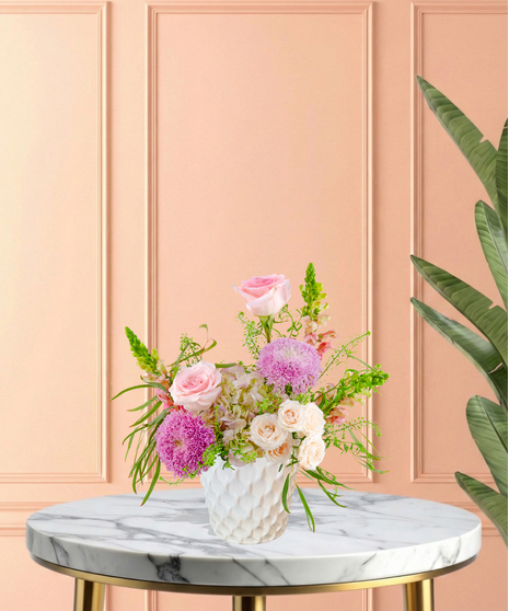 Lavender Sunrise floral arrangement with peach and blush roses, lavender pom blooms, snapdragons, and hydrangeas in a white ceramic vase.