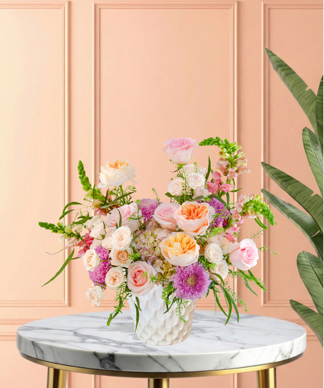 Lavender Sunrise floral arrangement with peach and blush roses, lavender pom blooms, snapdragons, and hydrangeas in a white ceramic vase.