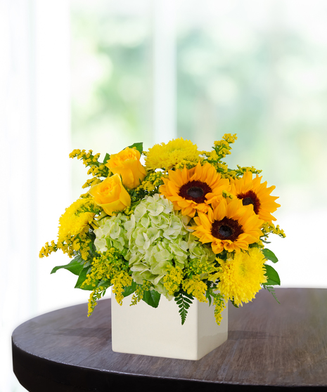 Lemon Drop floral arrangement with sunflowers, yellow roses, green hydrangeas, and solidago in a modern white vase on a wooden table.