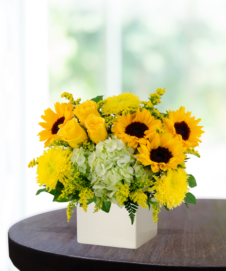 Lemon Drop floral arrangement with sunflowers, yellow roses, green hydrangeas, and solidago in a modern white vase on a wooden table.