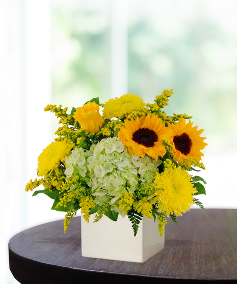 Lemon Drop floral arrangement with sunflowers, yellow roses, green hydrangeas, and solidago in a modern white vase on a wooden table.