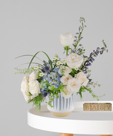 Little Lamb flower arrangement with white roses, blue hydrangea, delphinium, and chamomile in a striped ceramic vase for baby boy