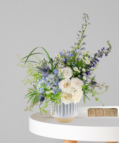 Little Lamb flower arrangement with white roses, blue hydrangea, delphinium, and chamomile in a striped ceramic vase for baby boy