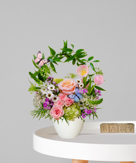 Lullaby Garden new baby flower arrangement with pink roses, tulips, daisy poms, and butterfly accents in a white ceramic vase