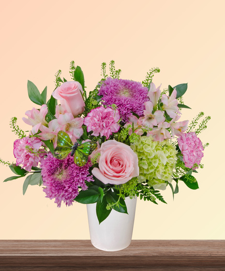 Made for Mama flower arrangement with pink roses, carnations, lavender blooms, green hydrangeas, and butterfly accent in a soft pink vase.