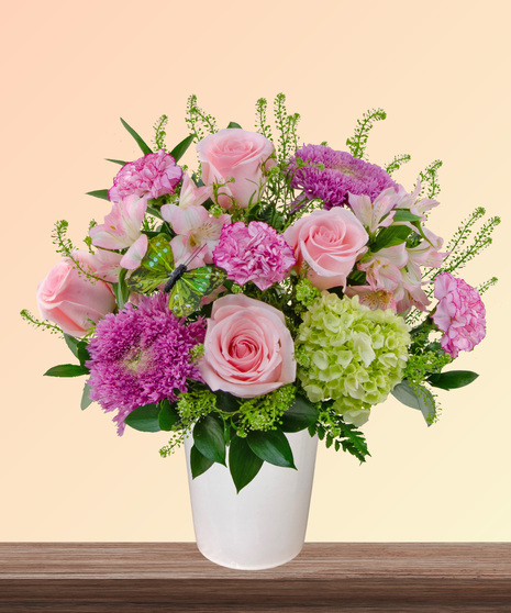 Made for Mama flower arrangement with pink roses, carnations, lavender blooms, green hydrangeas, and butterfly accent in a soft pink vase.
