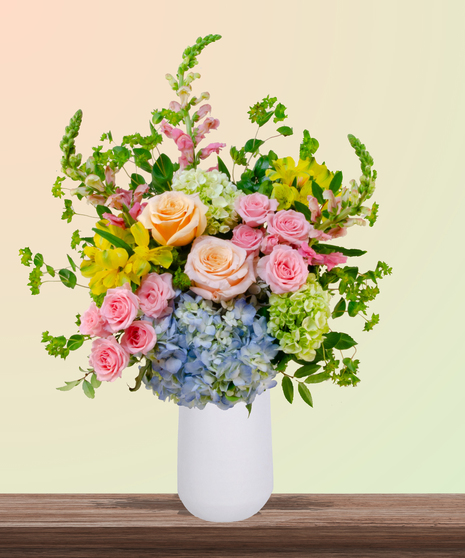 Because She’s Mom flower arrangement with peach and blush roses, blue hydrangeas, yellow alstroemeria, pink snapdragons, and greenery in a white vase.