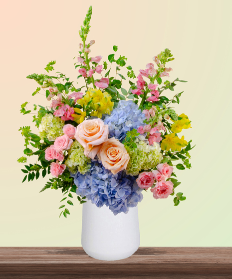 Because She’s Mom flower arrangement with peach and blush roses, blue hydrangeas, yellow alstroemeria, pink snapdragons, and greenery in a white vase.