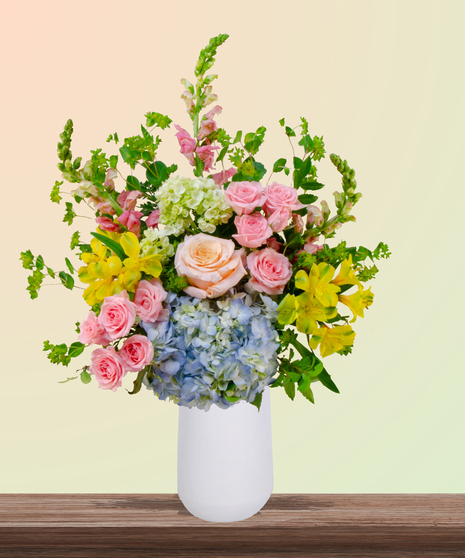 Because She’s Mom flower arrangement with peach and blush roses, blue hydrangeas, yellow alstroemeria, pink snapdragons, and greenery in a white vase.