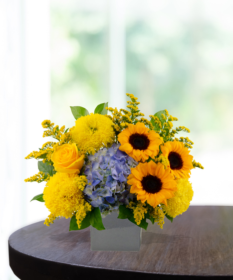 Main Squeeze floral arrangement featuring bright sunflowers, yellow roses, yellow pompon mums, and blue hydrangea in a gray ceramic vase