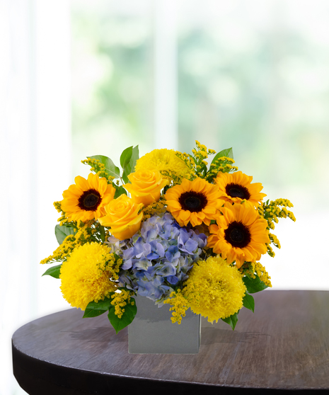 Main Squeeze floral arrangement featuring bright sunflowers, yellow roses, yellow pompon mums, and blue hydrangea in a gray ceramic vase