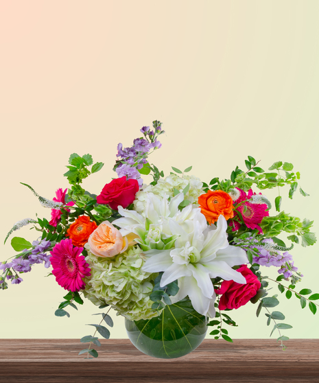 Mama Mia! floral arrangement with peach roses, white lilies, pink gerberas, orange ranunculus, green hydrangeas, and red roses in a round leaf-lined glass vase.