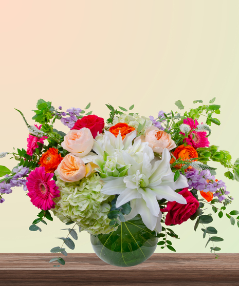 Mama Mia! floral arrangement with peach roses, white lilies, pink gerberas, orange ranunculus, green hydrangeas, and red roses in a round leaf-lined glass vase.