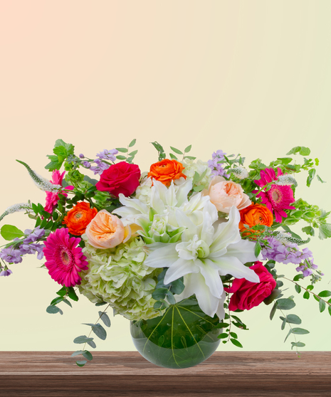 Mama Mia! floral arrangement with peach roses, white lilies, pink gerberas, orange ranunculus, green hydrangeas, and red roses in a round leaf-lined glass vase.