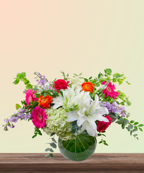 Mama Mia! floral arrangement with peach roses, white lilies, pink gerberas, orange ranunculus, green hydrangeas, and red roses in a round leaf-lined glass vase.