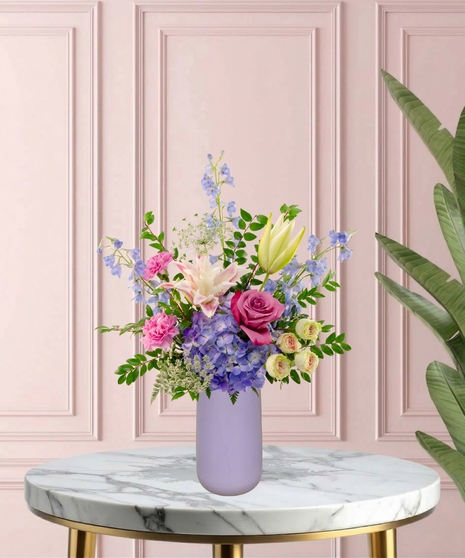Mama's Meadow Mother's Day flower arrangement featuring yellow garden roses, blue delphinium, purple hydrangeas, and pink-tipped rose lilies in a matte lavender vase.