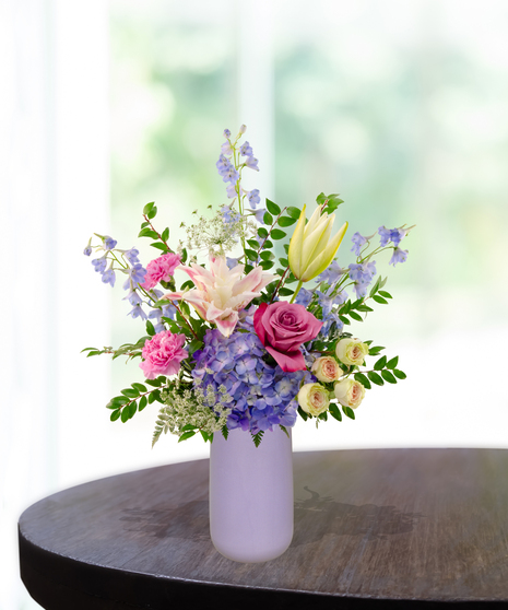 Mama's Meadow Mother's Day flower arrangement featuring yellow garden roses, blue delphinium, purple hydrangeas, and pink-tipped rose lilies in a matte lavender vase.