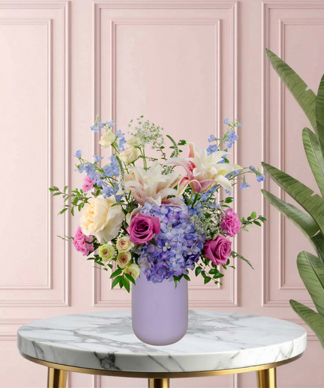 Mama's Meadow Mother's Day flower arrangement featuring yellow garden roses, blue delphinium, purple hydrangeas, and pink-tipped rose lilies in a matte lavender vase.