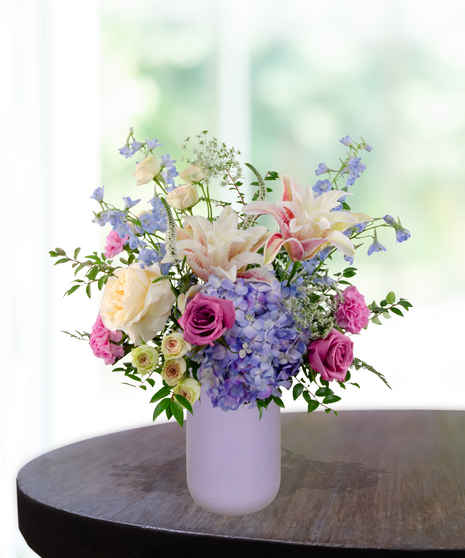 Mama's Meadow Mother's Day flower arrangement featuring yellow garden roses, blue delphinium, purple hydrangeas, and pink-tipped rose lilies in a matte lavender vase.