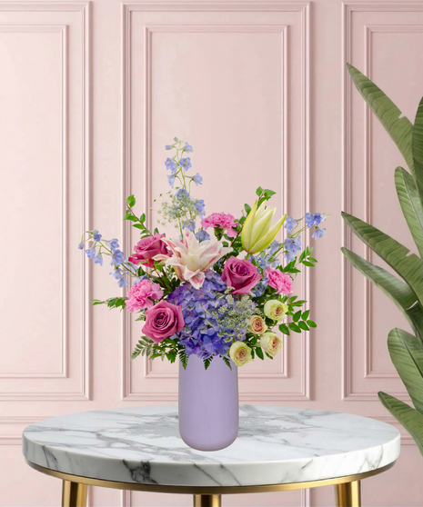 Mama's Meadow Mother's Day flower arrangement featuring yellow garden roses, blue delphinium, purple hydrangeas, and pink-tipped rose lilies in a matte lavender vase.