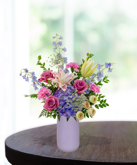 Mama's Meadow Mother's Day flower arrangement featuring yellow garden roses, blue delphinium, purple hydrangeas, and pink-tipped rose lilies in a matte lavender vase.