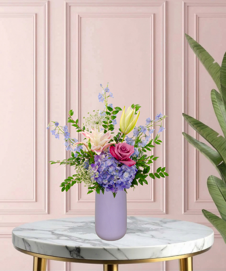 Mama's Meadow Mother's Day flower arrangement featuring yellow garden roses, blue delphinium, purple hydrangeas, and pink-tipped rose lilies in a matte lavender vase.