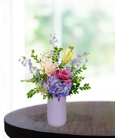 Mama's Meadow Mother's Day flower arrangement featuring yellow garden roses, blue delphinium, purple hydrangeas, and pink-tipped rose lilies in a matte lavender vase.