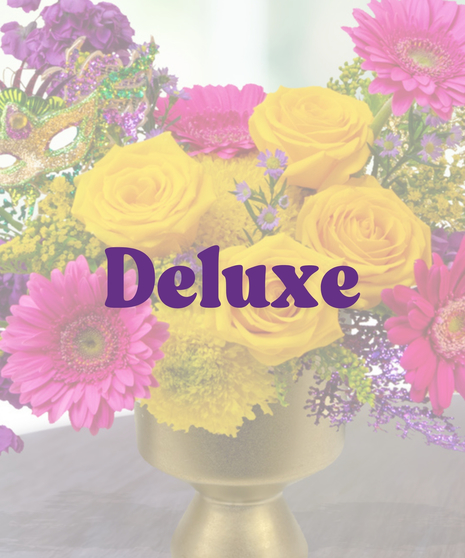 A Mardi Gras-themed arrangement featuring yellow roses, pink gerbera daisies, purple stock flowers, golden solidago, and a glittering mask in a gold pedestal vase.