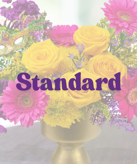 A Mardi Gras-themed arrangement featuring yellow roses, pink gerbera daisies, purple stock flowers, golden solidago, and a glittering mask in a gold pedestal vase.