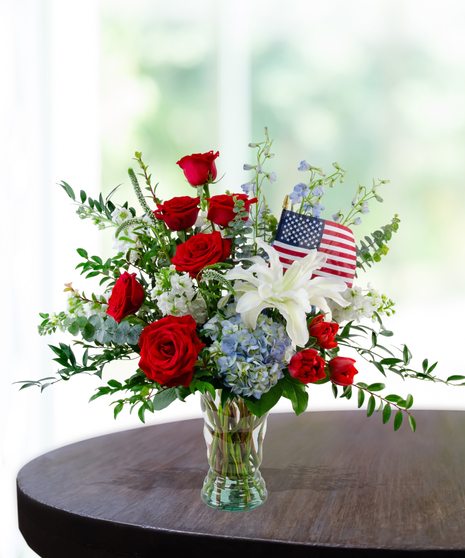 All-American patriotic flower arrangement featuring red roses, a white lily, blue hydrangeas, red tulips, and a small American flag in a clear glass vase.