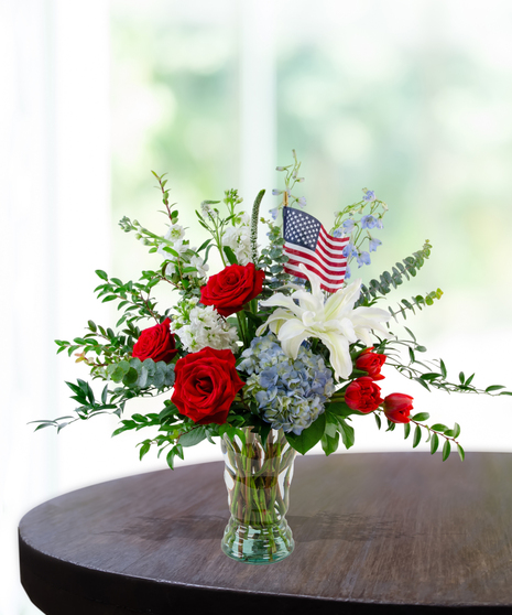 All-American patriotic flower arrangement featuring red roses, a white lily, blue hydrangeas, red tulips, and a small American flag in a clear glass vase.