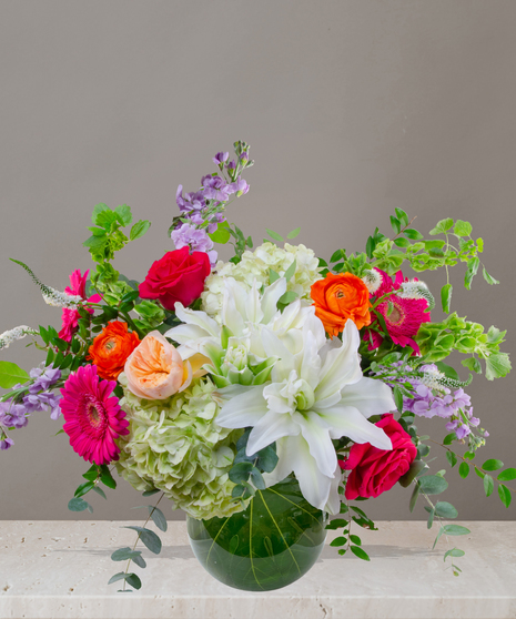 Mia flower arrangement with green hydrangeas, white lilies, pink gerberas, coral roses, and orange ranunculus in a tropical leaf-lined glass vase.