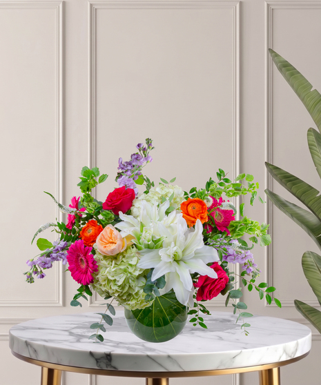 Mia flower arrangement with green hydrangeas, white lilies, pink gerberas, coral roses, and orange ranunculus in a tropical leaf-lined glass vase.