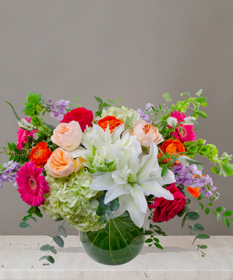 Mia flower arrangement with green hydrangeas, white lilies, pink gerberas, coral roses, and orange ranunculus in a tropical leaf-lined glass vase.