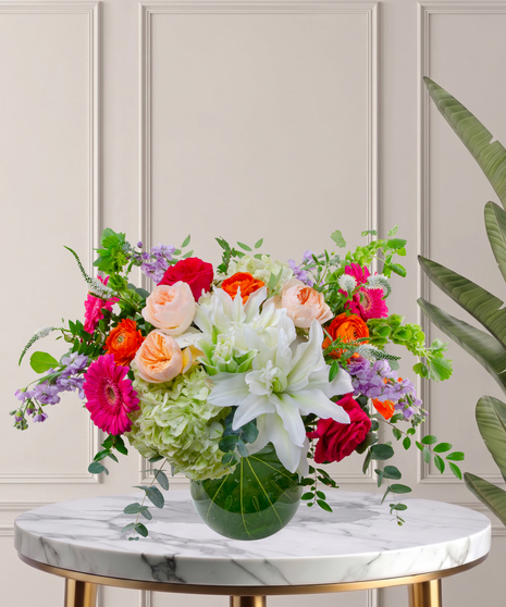 Mia flower arrangement with green hydrangeas, white lilies, pink gerberas, coral roses, and orange ranunculus in a tropical leaf-lined glass vase.