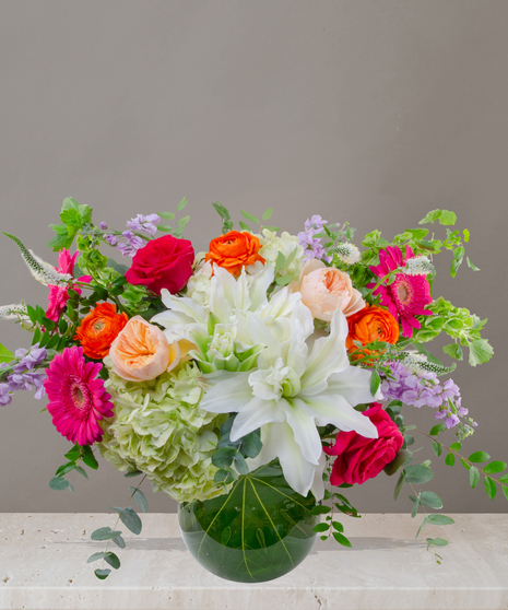Mia flower arrangement with green hydrangeas, white lilies, pink gerberas, coral roses, and orange ranunculus in a tropical leaf-lined glass vase.