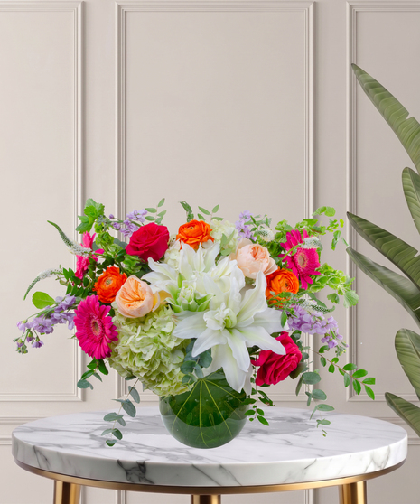 Mia flower arrangement with green hydrangeas, white lilies, pink gerberas, coral roses, and orange ranunculus in a tropical leaf-lined glass vase.