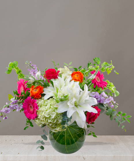 Mia flower arrangement with green hydrangeas, white lilies, pink gerberas, coral roses, and orange ranunculus in a tropical leaf-lined glass vase.