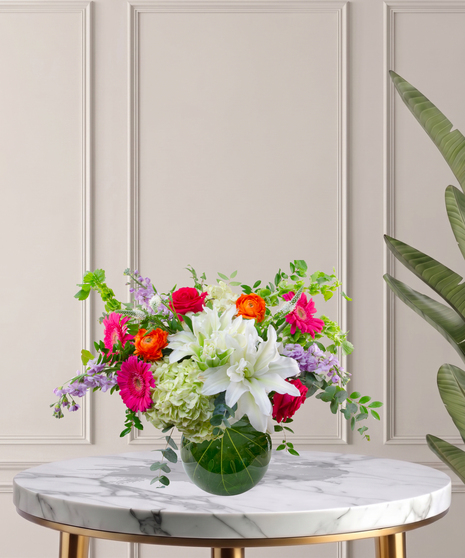 Mia flower arrangement with green hydrangeas, white lilies, pink gerberas, coral roses, and orange ranunculus in a tropical leaf-lined glass vase.
