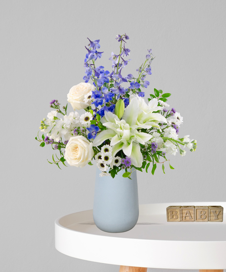 Misty Blues new baby flower arrangement featuring blue delphinium, white roses, white lilies, and blue hydrangeas in a soft blue ceramic vase next to wooden baby blocks