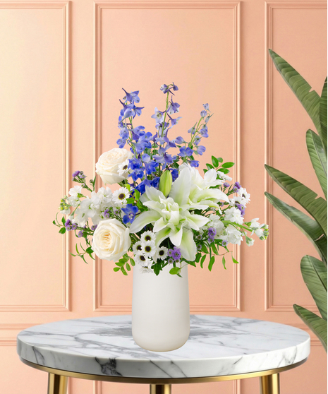 Gingham & Grace Easter flower arrangement with white roses, lilies, blue delphinium, blue hydrangeas, white snapdragons, purple accents, and foliage in a modern white vase