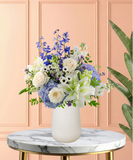Gingham & Grace Easter flower arrangement with white roses, lilies, blue delphinium, blue hydrangeas, white snapdragons, purple accents, and foliage in a modern white vase