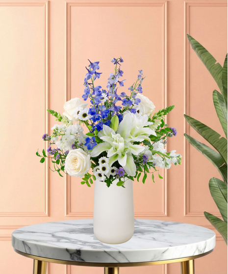 Gingham & Grace Easter flower arrangement with white roses, lilies, blue delphinium, blue hydrangeas, white snapdragons, purple accents, and foliage in a modern white vase