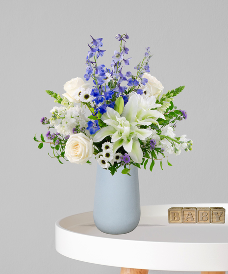 Misty Blues new baby flower arrangement featuring blue delphinium, white roses, white lilies, and blue hydrangeas in a soft blue ceramic vase next to wooden baby blocks