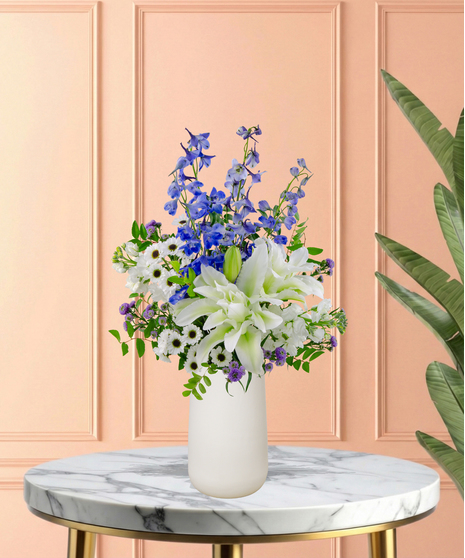 Gingham & Grace Easter flower arrangement with white roses, lilies, blue delphinium, blue hydrangeas, white snapdragons, purple accents, and foliage in a modern white vase