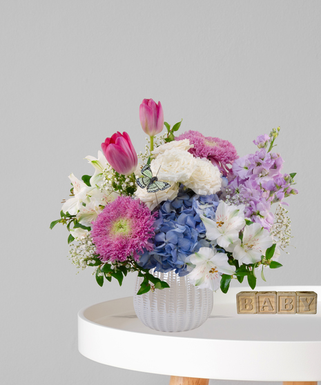 Little Moonbeam flower arrangement with white roses, blue hydrangeas, pink tulips, and butterfly accent in a white vase for new baby gift