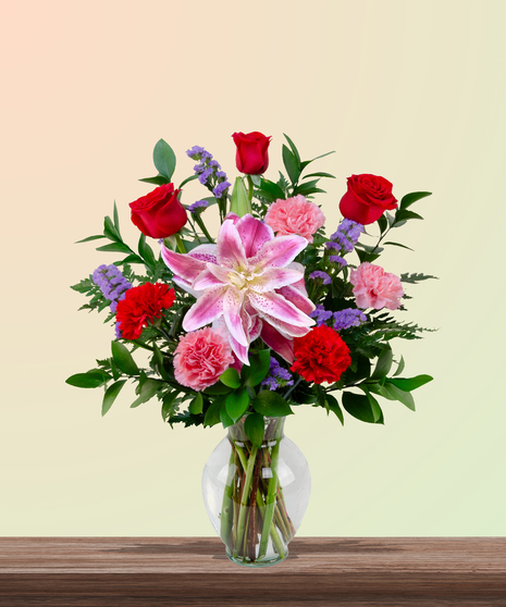Mother’s Touch floral arrangement with red roses, pink carnations, stargazer lilies, and purple statice in a clear glass vase – a classic Mother’s Day gift.