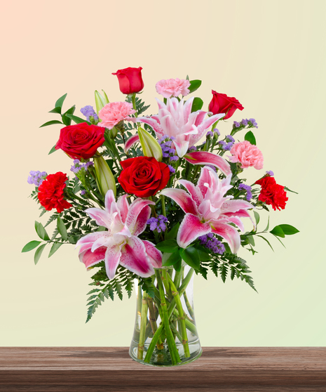Mother’s Touch floral arrangement with red roses, pink carnations, stargazer lilies, and purple statice in a clear glass vase – a classic Mother’s Day gift.
